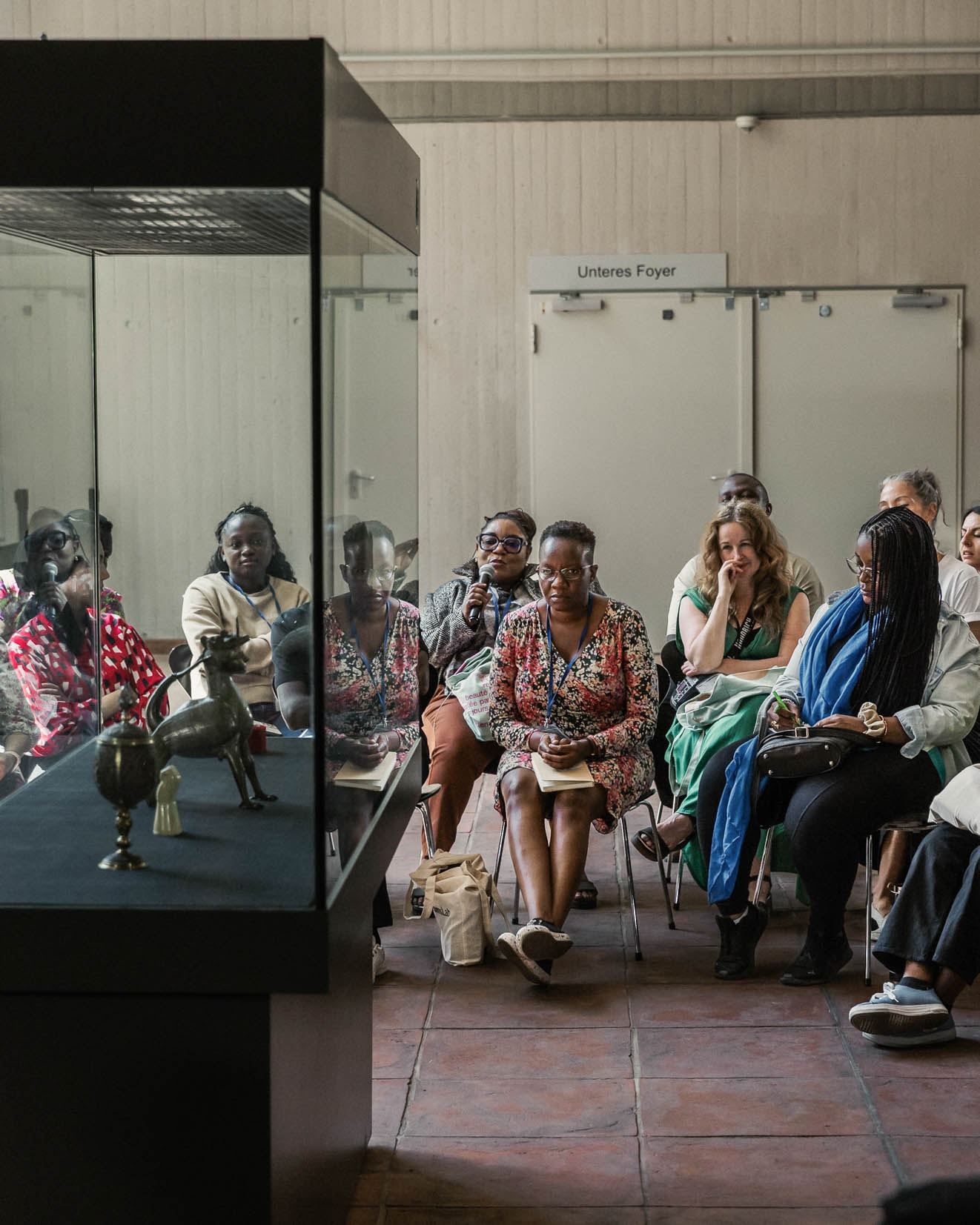 Fellows examine objects in a display case while one Fellow speaks into a microphone.